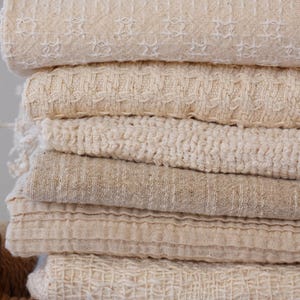 May include: A pile of folded, cream-coloured textiles with diverse textures. The fabrics feature woven, ribbed, and patterned designs. The stack is placed on a woven, light brown surface, indicating a home decor or textile product.