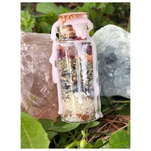May include: A small glass bottle filled with dried herbs, flowers, and crystals, sealed with a cork and topped with a pink wax seal. The bottle is surrounded by green foliage and rocks.