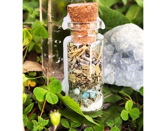 May include: A small glass bottle filled with dried herbs, crystals, and other natural elements. The bottle has a cork stopper and is surrounded by green foliage.