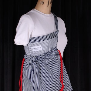 May include: A white short-sleeved shirt is worn under a blue and white striped apron-style garment with red drawstrings. The apron has a label that reads "ROBINSON HERITAGE WEAR". The garment is displayed on a mannequin.