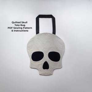 Può includere: Una borsa tote a forma di teschio trapuntato, grigio chiaro, con manico nero. Il teschio ha orbite e naso neri. Il testo "Quilted Skull Tote Bag PDF Sewing Pattern & Instructions" è visibile.