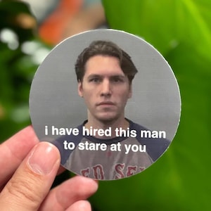 May include: A circular sticker featuring a photograph of a person with dark hair wearing a grey and navy blue baseball-style shirt. The text "i have hired this man to stare at you" is printed in white letters across the center of the sticker.