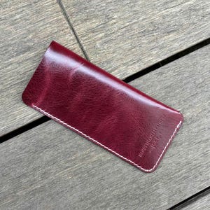 May include: A deep burgundy leather sleeve with a smooth, glossy finish and visible white stitching along the edges. The surface features a subtle grain texture and includes the embossed text "HANDSTITCHED LEATHER" in the bottom right corner.