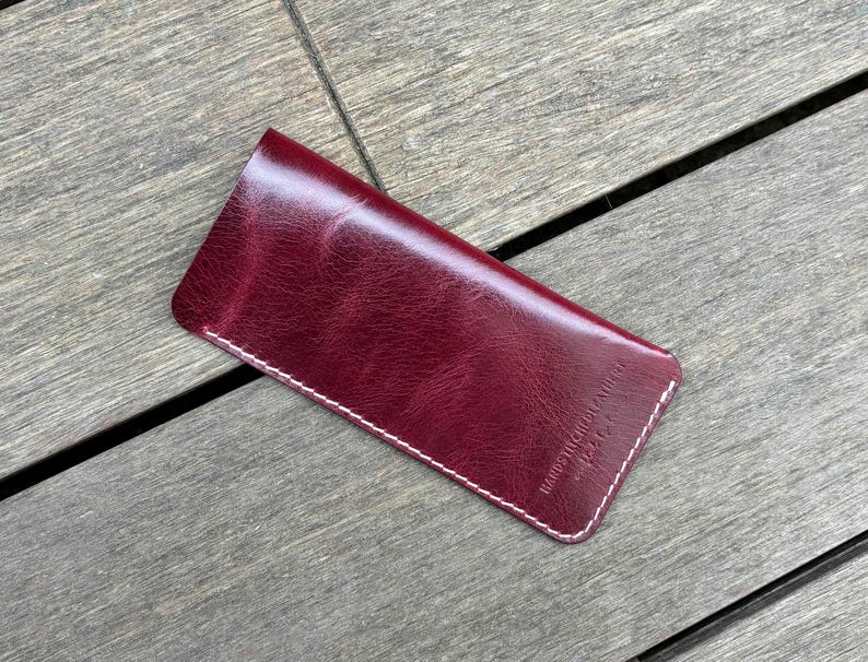 May include: A deep burgundy leather sleeve with a smooth, glossy finish and visible white stitching along the edges. The surface features a subtle grain texture and includes the embossed text "HANDSTITCHED LEATHER" in the bottom right corner.