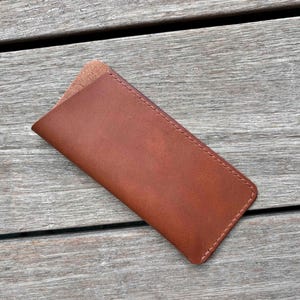 May include: A rectangular brown leather sleeve with rounded corners and visible stitching along the edges. The case features a smooth exterior surface and a slightly textured, lighter brown interior lining visible at the top opening of the pouch.