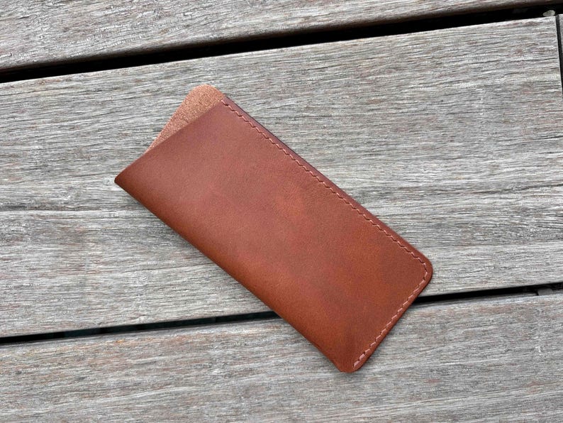 May include: A rectangular brown leather sleeve with rounded corners and visible stitching along the edges. The case features a smooth exterior surface and a slightly textured, lighter brown interior lining visible at the top opening of the pouch.