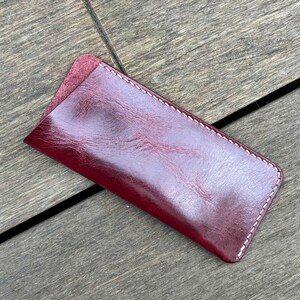 May include: A rectangular burgundy leather sleeve with rounded corners and white contrast stitching along the edges. The surface of the leather has a smooth, polished finish with visible natural grain patterns and a slightly darker, textured opening.
