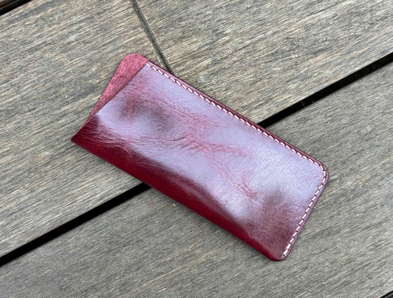 May include: A rectangular burgundy leather sleeve with rounded corners and white contrast stitching along the edges. The surface of the leather has a smooth, polished finish with visible natural grain patterns and a slightly darker, textured opening.