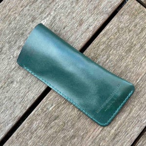 May include: A forest green leather sleeve case with a smooth finish and tonal stitching along the edges. The surface features a small embossed logo that reads "KINDSTITCHLEATHER" in a clean, sans-serif font, positioned near the bottom right corner.