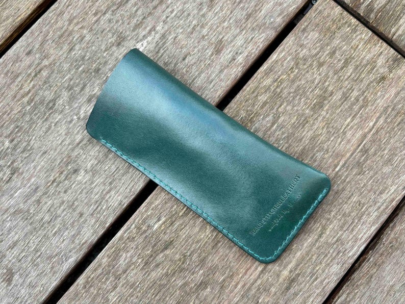May include: A forest green leather sleeve case with a smooth finish and tonal stitching along the edges. The surface features a small embossed logo that reads "KINDSTITCHLEATHER" in a clean, sans-serif font, positioned near the bottom right corner.