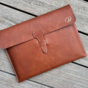 May include: A tan leather document sleeve featuring a fold-over flap with a secure strap closure. The exterior is accented with white contrast stitching along the edges and a small, elegant letter D monogram embossed in the upper right corner.