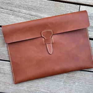 May include: A rectangular brown leather document case featuring a fold-over flap closure secured by a central leather strap and loop mechanism. The edges are finished with visible white stitching that outlines the perimeter and the closure.