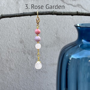 May include: A decorative charm labeled 3. Rose Garden featuring a gold-tone metal lobster clasp, a vertical arrangement of three round beads in pink, lavender, and white, and a single white teardrop-shaped bead suspended at the bottom by gold wire.