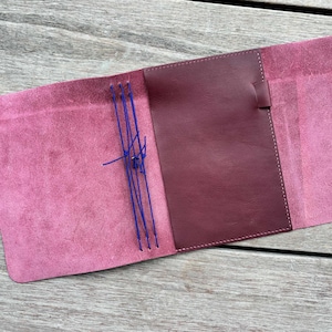 May include: A magenta leather notebook cover featuring a smooth dark burgundy interior pocket and three vertical blue elastic cords for securing inserts. The cover has a soft, suede-like texture on the outer panels and a pen loop.