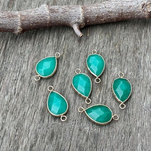 May include: Six teal-coloured, teardrop-shaped gemstone pendants with gold-coloured metal edging and loops. The pendants are faceted. They are arranged on a weathered, grey wooden surface, with a small, brown twig in the background.