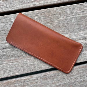 May include: A rectangular brown leather sleeve with rounded corners and visible light-colored stitching along the edges. The item features a smooth, matte finish and a simple, flat construction designed to hold small, thin objects securely.