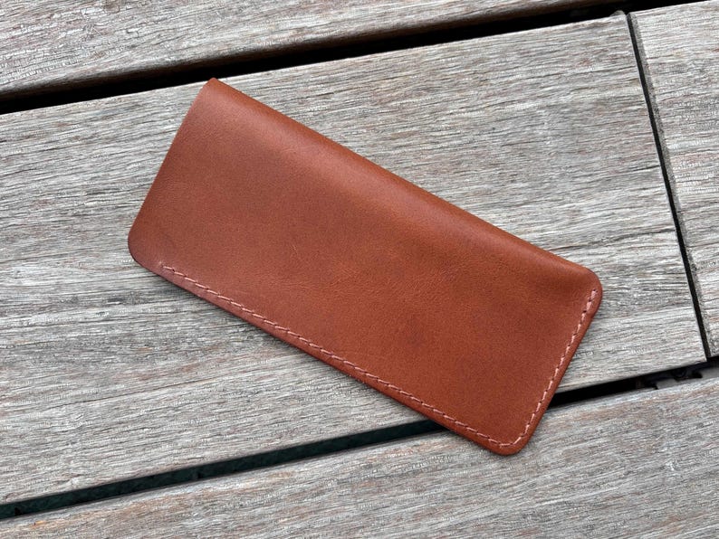 May include: A rectangular brown leather sleeve with rounded corners and visible light-colored stitching along the edges. The item features a smooth, matte finish and a simple, flat construction designed to hold small, thin objects securely.