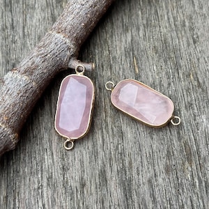 May include: Two rose quartz gemstone connectors with gold-colored bezels. The gemstones are rectangular with faceted surfaces, displaying a soft pink hue. Each connector has a loop at both ends for attaching to other jewelry components. The background is a weathered wooden surface.