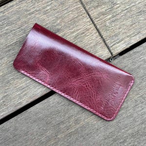May include: A rectangular burgundy leather case with a glossy, textured finish and rounded corners. The item features visible stitching along the edges and the embossed text HAND STITCHED LEATHER on the lower right corner of the front surface.
