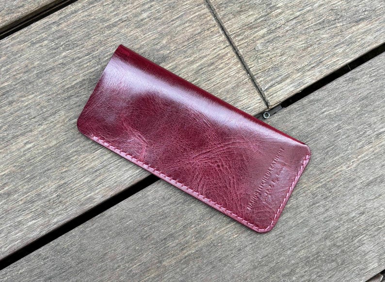 May include: A rectangular burgundy leather case with a glossy, textured finish and rounded corners. The item features visible stitching along the edges and the embossed text HAND STITCHED LEATHER on the lower right corner of the front surface.