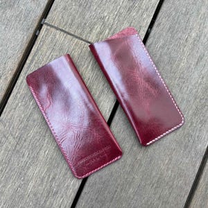May include: Two rectangular burgundy leather sleeves with rounded bottom corners and white contrast stitching along the edges. The leather has a glossy, textured finish with visible grain patterns. One sleeve features embossed text near the bottom.