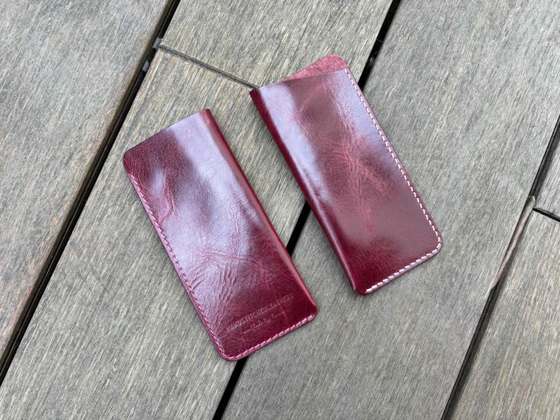 May include: Two rectangular burgundy leather sleeves with rounded bottom corners and white contrast stitching along the edges. The leather has a glossy, textured finish with visible grain patterns. One sleeve features embossed text near the bottom.