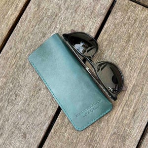May include: A teal leather glasses case with visible stitching along the edges and the embossed text HANDSTITCHEDLEATHERY crafted by lucas, paired with a pair of black-framed sunglasses with dark lenses resting on top of the case.