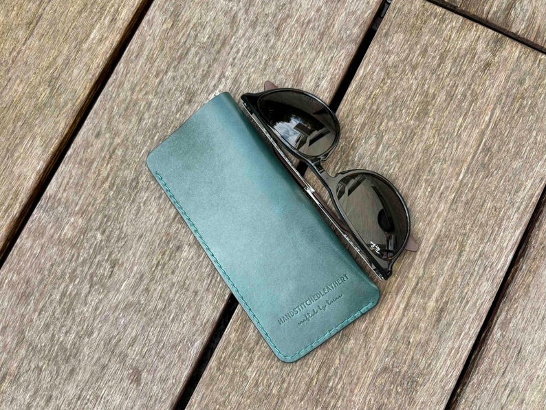 May include: A teal leather glasses case with visible stitching along the edges and the embossed text HANDSTITCHEDLEATHERY crafted by lucas, paired with a pair of black-framed sunglasses with dark lenses resting on top of the case.