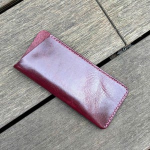May include: A rectangular burgundy leather case with a smooth, polished finish and a diagonal opening. The edges are secured with matching burgundy stitching, creating a clean, structured silhouette for holding small items like glasses or pens.