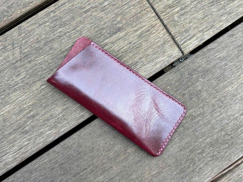 May include: A rectangular burgundy leather case with a smooth, polished finish and a diagonal opening. The edges are secured with matching burgundy stitching, creating a clean, structured silhouette for holding small items like glasses or pens.