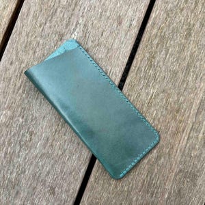 May include: A rectangular teal leather sleeve with rounded bottom corners and visible tonal stitching along the edges. The sleeve features a diagonal opening at the top, designed to hold small items securely within its smooth, flat construction.