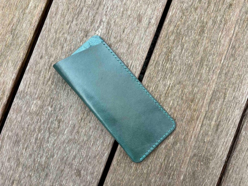 May include: A rectangular teal leather sleeve with rounded bottom corners and visible tonal stitching along the edges. The sleeve features a diagonal opening at the top, designed to hold small items securely within its smooth, flat construction.
