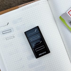 Könnte beinhalten: Ein schwarzer Haftnotizspender mit dem Aufdruck "allemore", der verschiedene Band- und Bannerdesigns aufweist. Der Spender befindet sich auf einem offenen Notizbuch mit kariertem Papier. Im Hintergrund befindet sich eine Packung mit einem Stift.