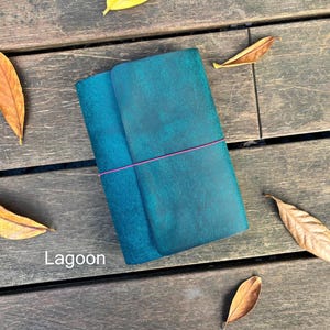 Op de afbeelding: Een rechthoekig blauwgroen lederen notitieboek met een textuurafwerking, vastgezet met een dun roze elastiek dat horizontaal om de kaft is gewikkeld. Het woord Lagoon staat in witte letters in de linkerbenedenhoek van de kaft gedrukt.