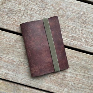 May include: A rectangular notebook with a dark brown, textured cover featuring a mottled appearance. A single, vertical, light olive green elastic band is wrapped around the center of the notebook to keep the cover securely closed.