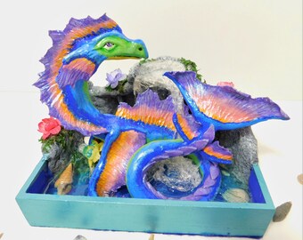 Könnte beinhalten: Eine blaue und violette Drachen-Skulptur mit orangefarbenen und rosafarbenen Akzenten, die in einem blauen und grünen, mit Wasser gefüllten Pflanztopf mit Muscheln und Blumen sitzt.