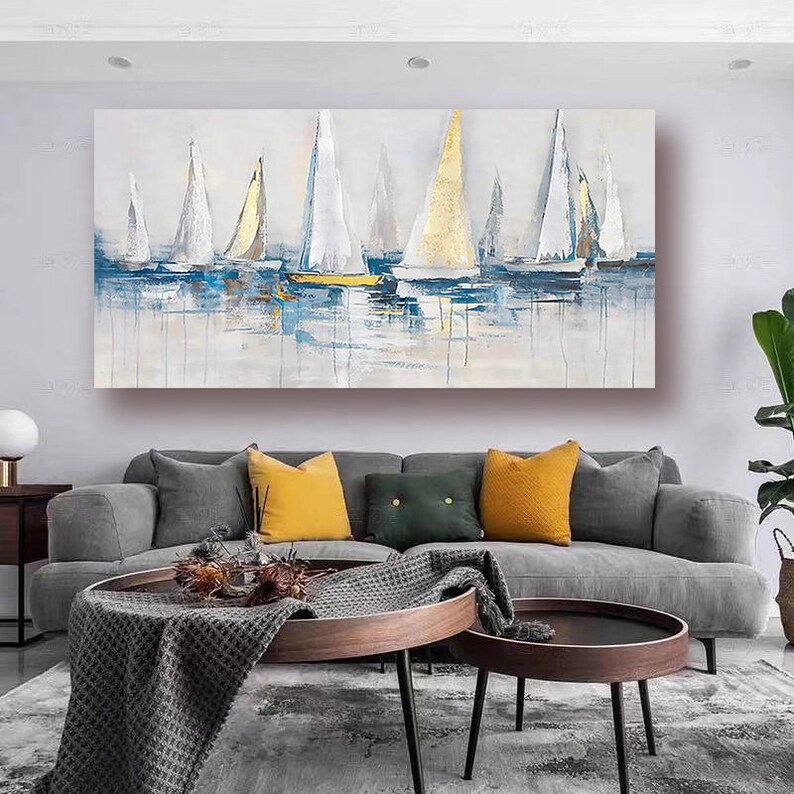 May include: Abstract painting of sailboats on a blue and white water background. The sailboats are white with gold accents. The painting is framed and hanging on a wall in a living room.
