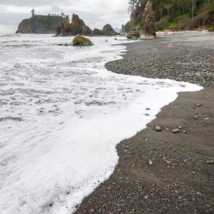 May include: A rocky beach with white foamy waves crashing on the shore. The beach is covered in small, dark pebbles. A line of trees is visible in the background.