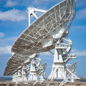 May include: A row of large white satellite dishes against a blue sky with white clouds. The dishes are arranged in a line and are all pointing in the same direction.