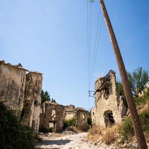 May include: An image of a deserted, sunlit street lined with the ruins of stone buildings. A wooden utility pole with glass insulators stands in the foreground, with power lines stretching across a clear blue sky.