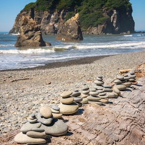 May include: A rocky beach with a large rock formation in the background. A wooden log sits on the beach with several small rock stacks balanced on top.