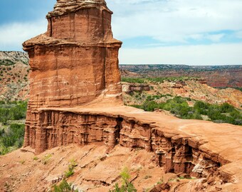 May include: A tall, red rock formation with a pointed top rises from a red, rocky landscape. The rock formation is surrounded by green trees and bushes.