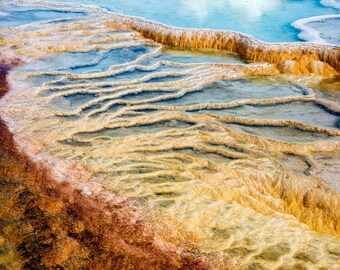 May include: Aerial view of a natural landscape featuring vibrant colors. Turquoise and blue water pools are surrounded by textured, golden-brown formations. The scene evokes a sense of geothermal activity and natural beauty.