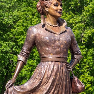 May include: Bronze statue of a woman in a polka-dot dress, holding a purse. The statue has a detailed hairstyle and a pearl necklace. The background is a blur of green trees.