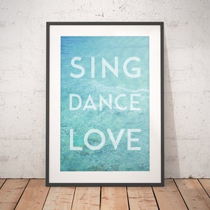 May include: Framed art print featuring the words "SING DANCE LOVE" in white capital letters against a blue and turquoise water background. The artwork is in a black frame and is displayed on a wooden floor.