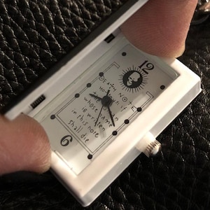Peut inclure: Une montre rectangulaire blanche avec un couvercle à charnière. Le cadran de la montre présente le texte "DEATH NOTE" et une illustration de crâne. Les aiguilles de la montre indiquent les chiffres 6 et 12. La montre est en cours d'ouverture.