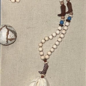 Puede incluir: Un collar de cuentas color crema con dijes de botas de vaquero marrones, cuentas de acento azules y marrones y una borla color crema. El collar está hecho de cuentas de madera y se muestra sobre un fondo neutro.
