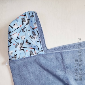 May include: A blue hooded towel with a blue and white patterned lining featuring hockey sticks, pucks, and skates. The towel is made of soft, absorbent material.