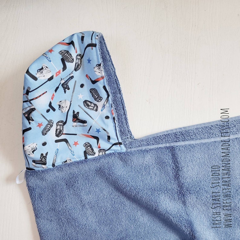 May include: A blue hooded towel with a blue and white patterned lining featuring hockey sticks, pucks, and skates. The towel is made of soft, absorbent material.