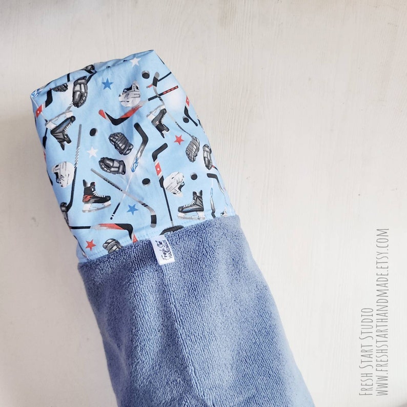 May include: A blue hooded towel with a hockey-themed print featuring hockey sticks, pucks, skates, and gloves. The towel has a blue terry cloth lining.
